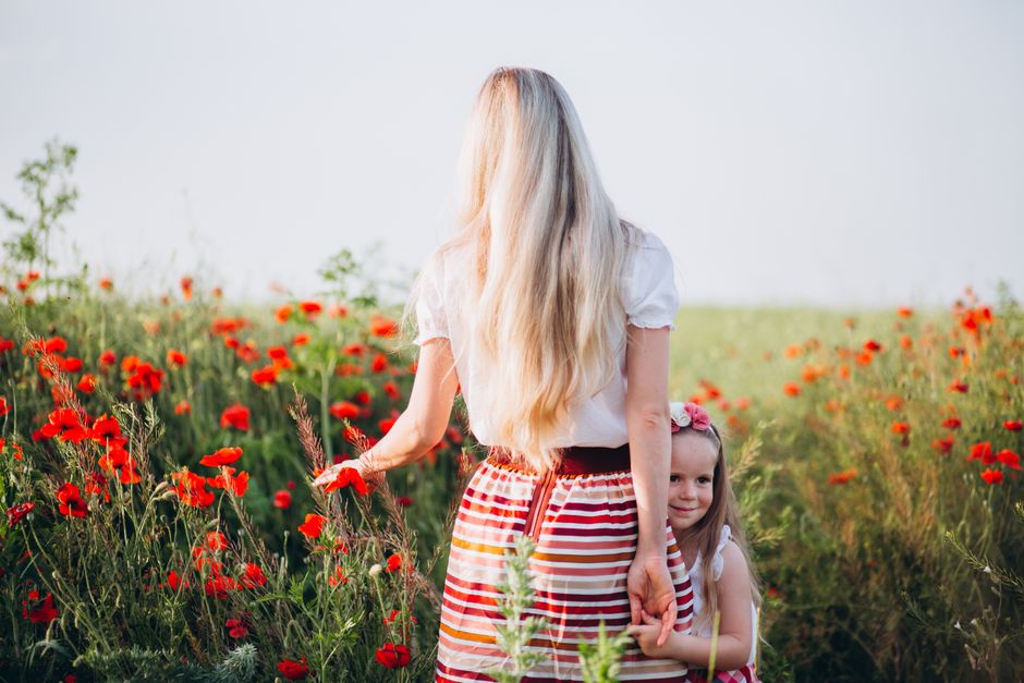 Happy little daughter in casual clothing looking away with smile while holding hand of faceless slim blond haired mother in field of poppies under cloudless sky